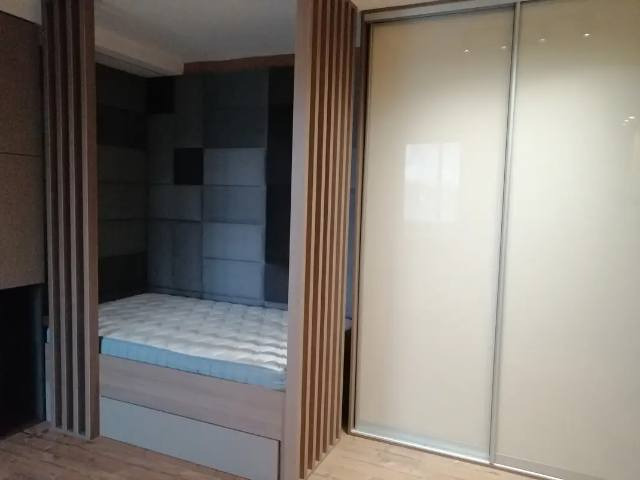 Wnęka z łóżkiem i szafa przesuwna z matowego szkła, drewniane panele dekoracyjne po bokach, szara tapicerowana ściana we wnęce.