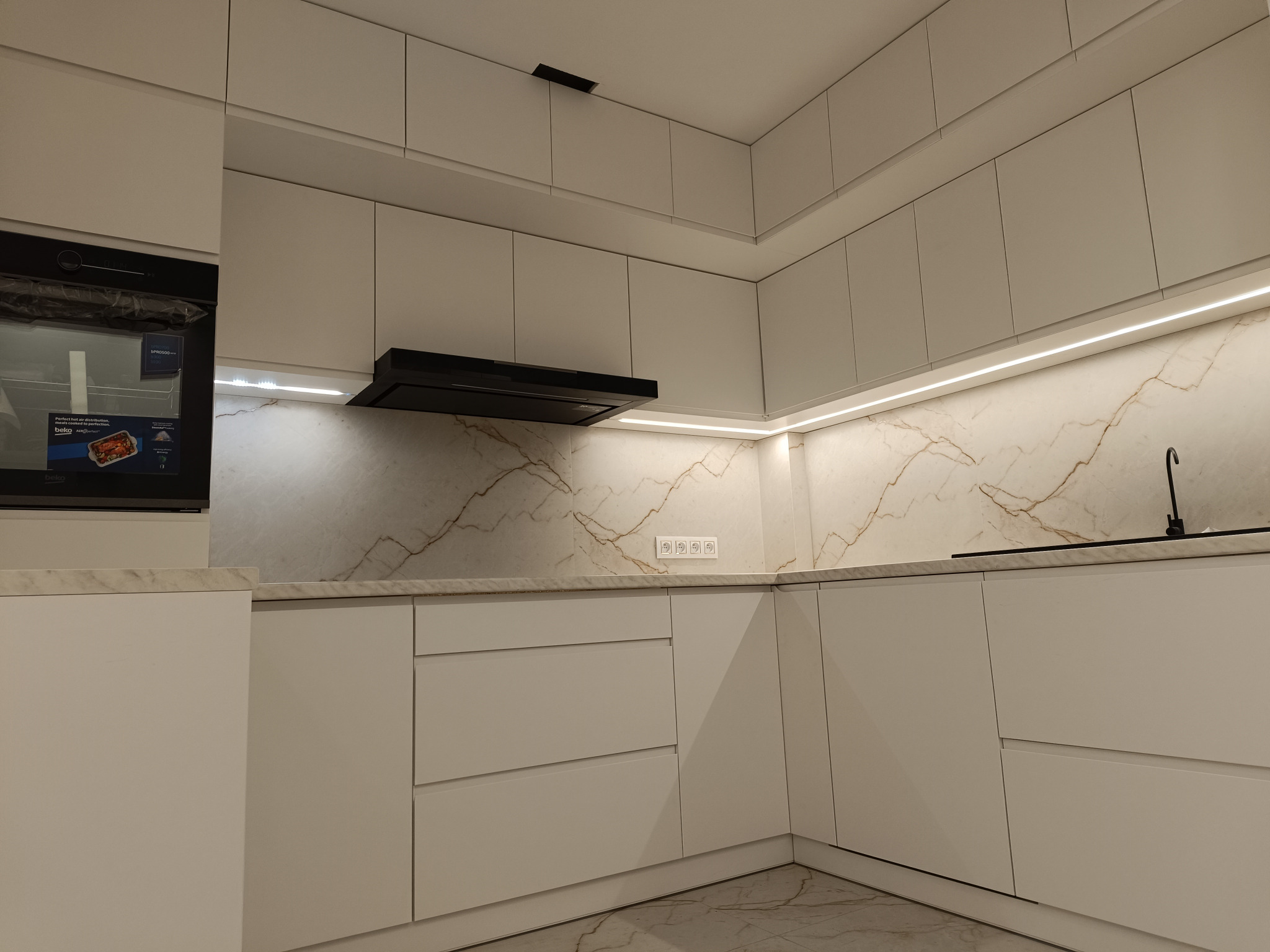 Nowoczesna, minimalistyczna kuchnia z białymi, gładkimi frontami szafek, marmurowym blatem i podświetleniem LED pod szafkami wiszącymi, widoczny piekarnik marki Beko i czarny okap