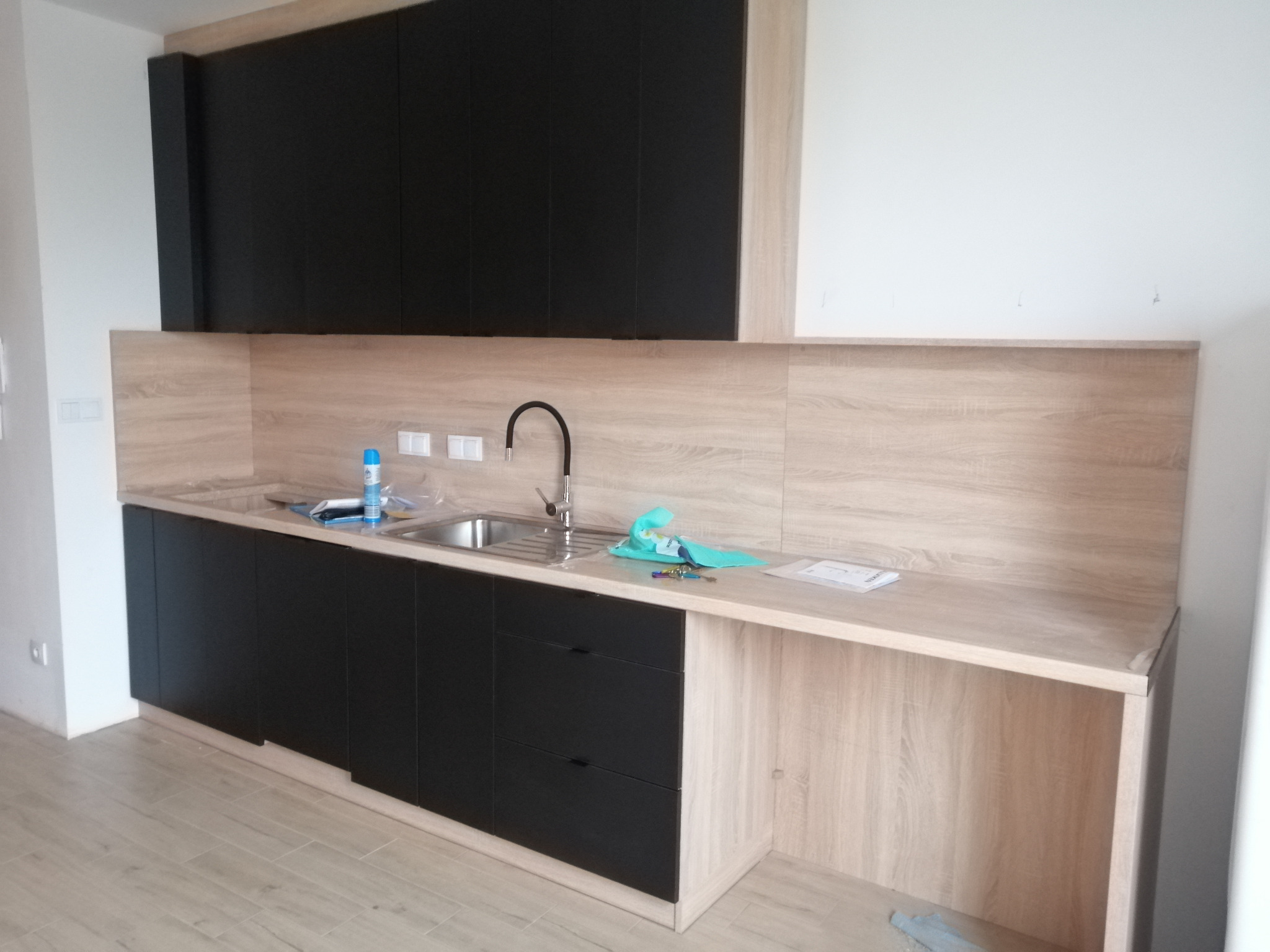 Nowoczesna kuchnia z czarnymi szafkami górnymi i dolnymi, blatem w kolorze jasnego drewna. Widoczny zlew, bateria i akcesoria kuchenne. Minimalistyczny design.