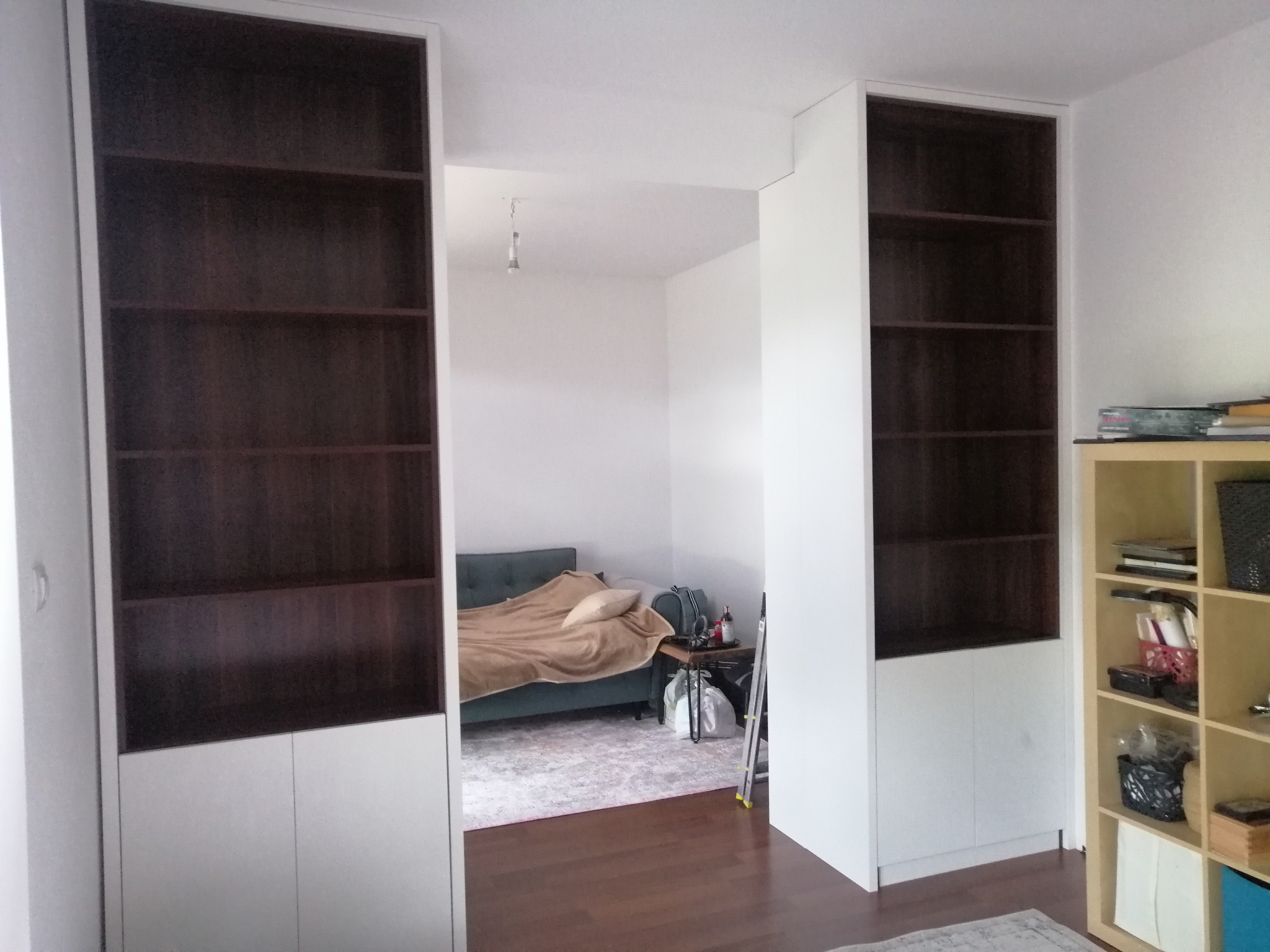 Białe szafy wnękowe z ciemnymi półkami, tworzące przejście do pokoju z kanapą. Minimalistyczny design, widoczne detale wykończenia i oświetlenie wnętrza.