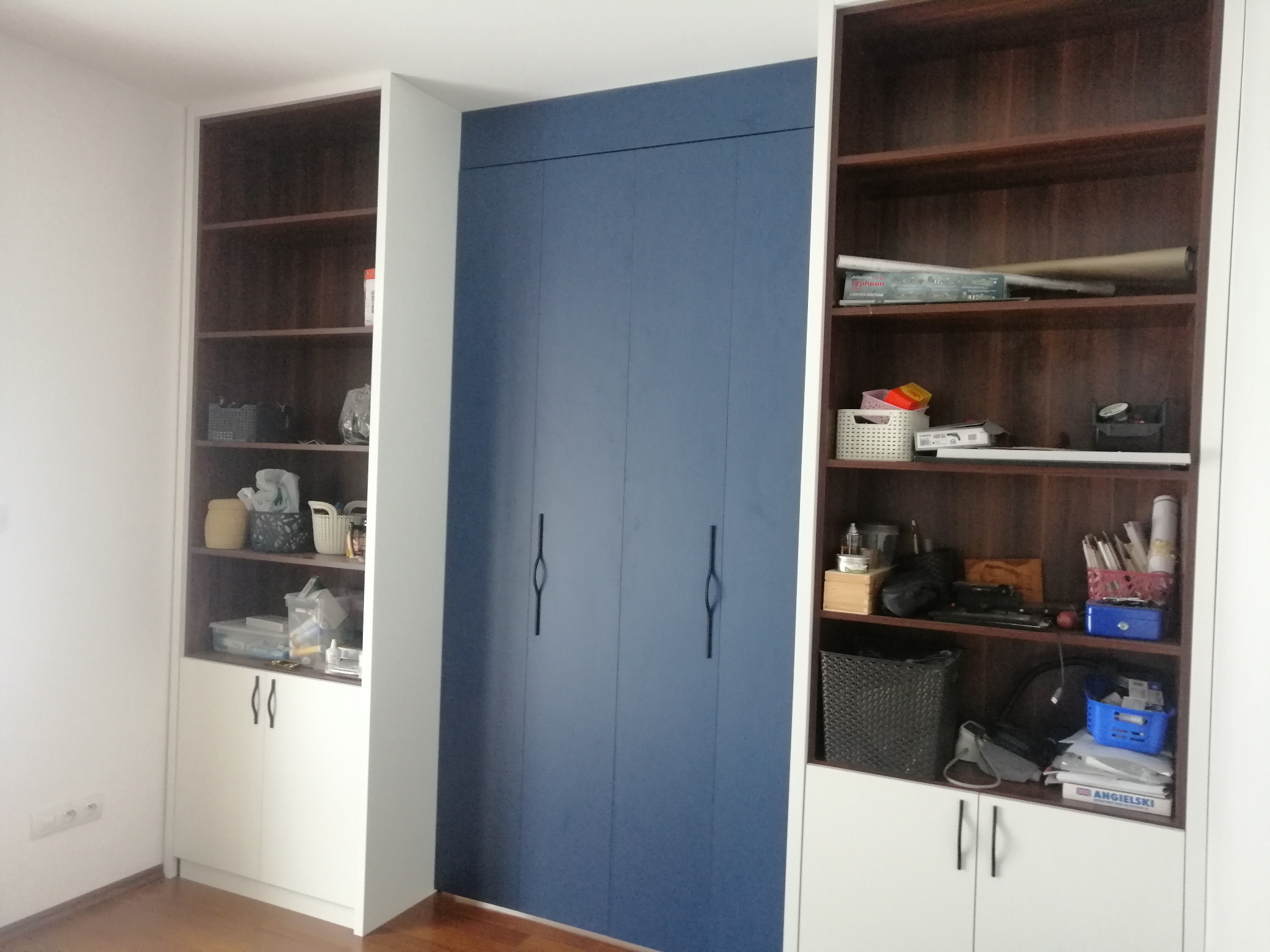 Zabudowa wnęki z szafą na wymiar i regałami po bokach. Szafa w kolorze granatowym, regały z ciemnego drewna z białymi dolnymi szafkami. Minimalistyczne uchwyty.