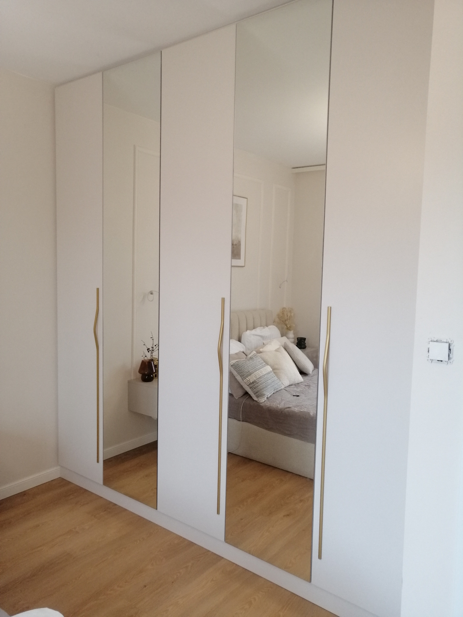 Biała szafa wnękowa z lustrzanymi drzwiami, widoczne złote uchwyty i odbicie sypialni. Minimalistyczny design, jasne wnętrze.
