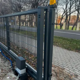 Szara, przesuwna brama wjazdowa z automatyką i żółtą lampą ostrzegawczą, widoczny silnik bramy i niebieski kabel zasilający, jesienne liście na ziemi.