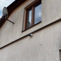 STREFA MONITORINGU - Elewacja domu z zamontowaną anteną satelitarną i kamerą monitoringu pod oknem, widoczne okablowanie.