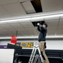 Pracownik na drabinie w sklepie, instalujący lub naprawiający okablowanie w suficie podwieszanym, z widocznym otworem rewizyjnym i elementami oświetlenia.