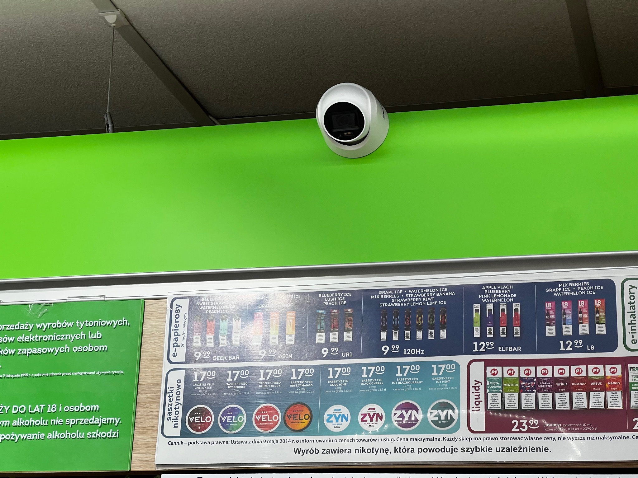 Biała kamera kopułkowa zamontowana na zielonej ścianie w sklepie nad regałami z e-papierosami i saszetkami nikotynowymi.