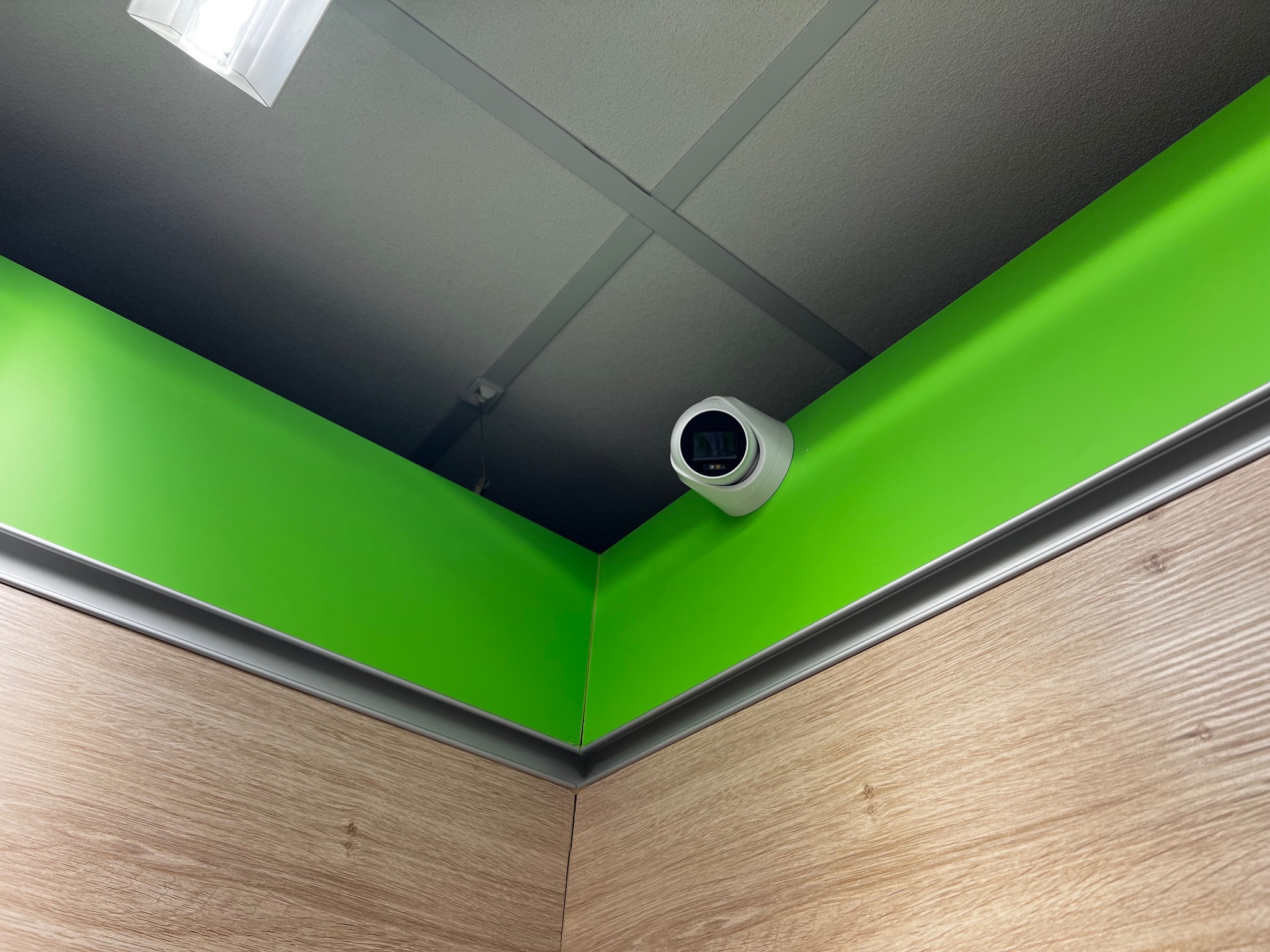 Kamera przemysłowa w rogu pomieszczenia z zielonymi ścianami i panelami drewnopodobnymi, widok z dołu na sufit z oświetleniem.