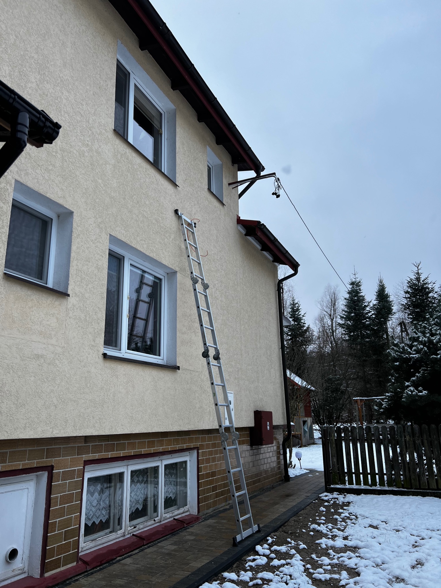 Drabina oparta o ścianę domu z zamontowaną anteną satelitarną na tle zimowego krajobrazu.