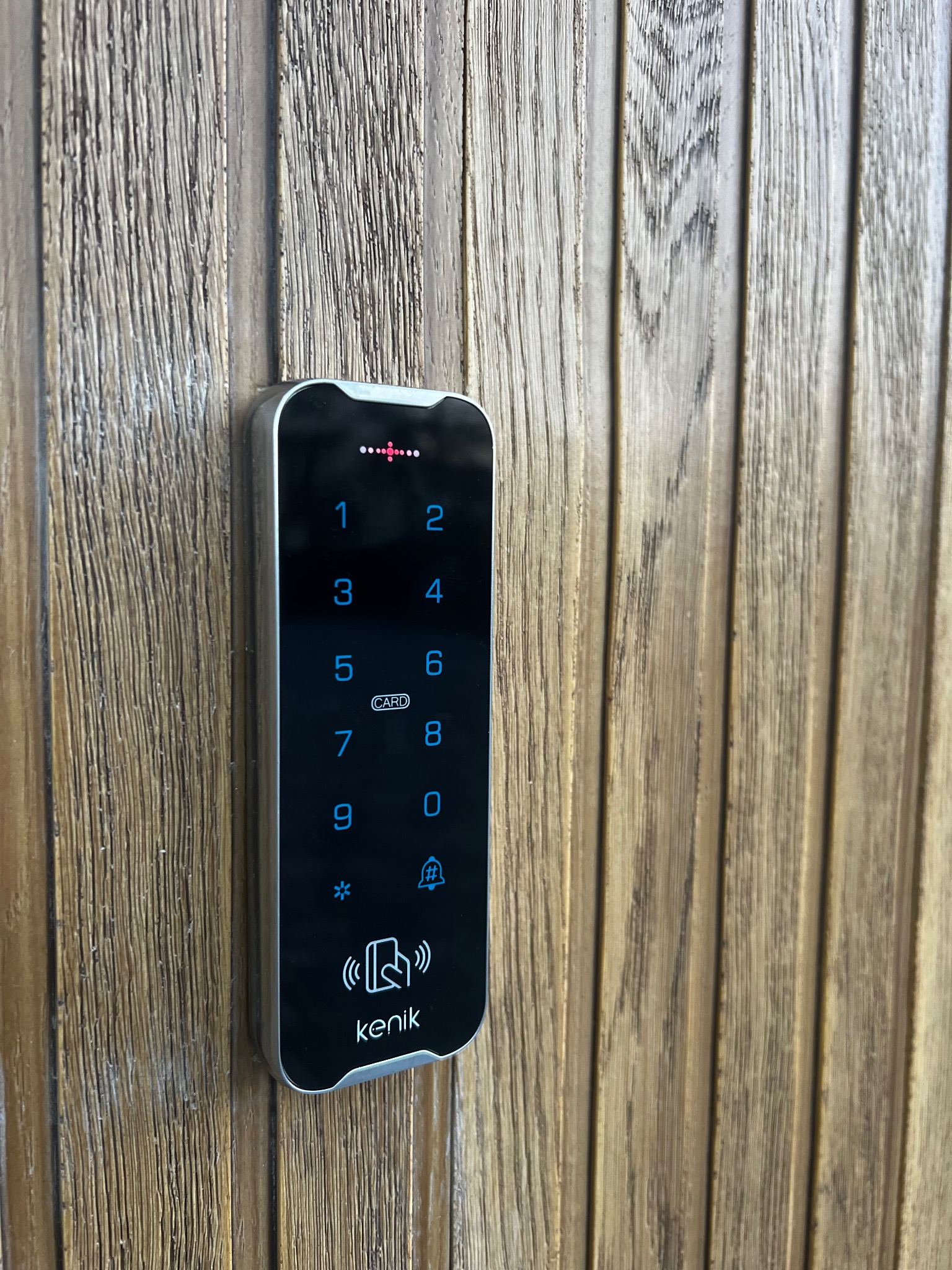 Elektroniczny zamek szyfrowy z czytnikiem kart zbliżeniowych, marki Kenik, zamontowany na drewnianych drzwiach z pionowymi panelami.