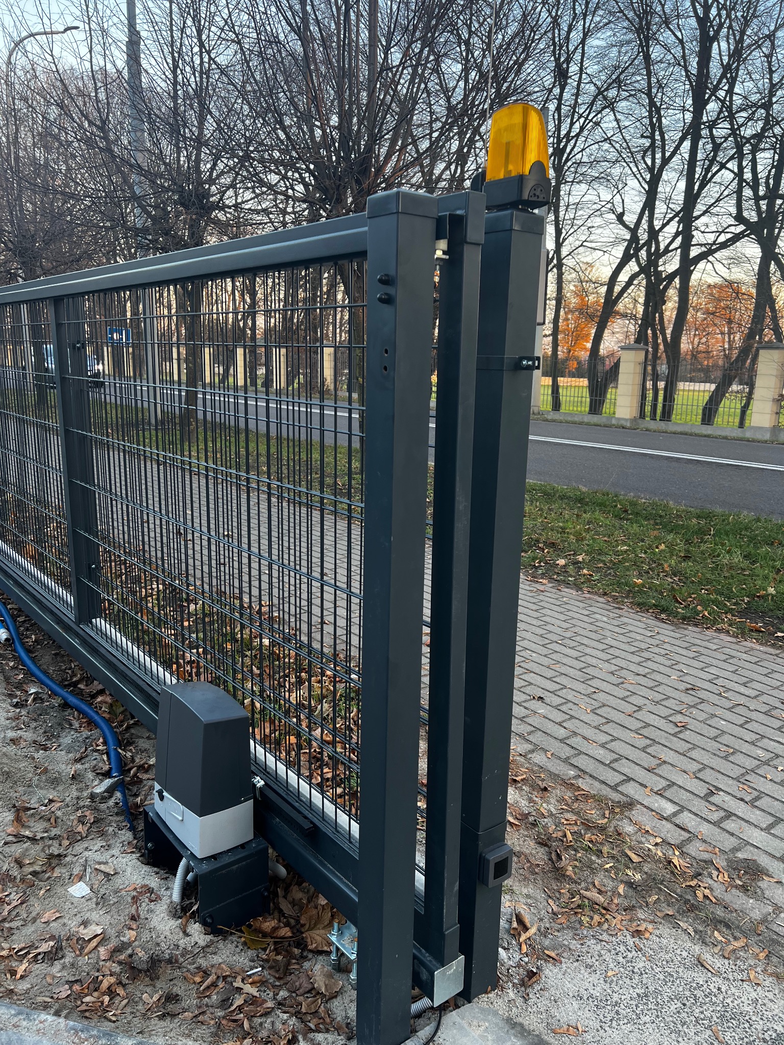 Szara, przesuwna brama wjazdowa z automatyką i żółtą lampą ostrzegawczą, widoczny silnik bramy i niebieski kabel zasilający, jesienne liście na ziemi.