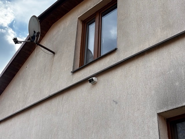 Elewacja domu z zamontowaną anteną satelitarną i kamerą monitoringu pod oknem, widoczne okablowanie.