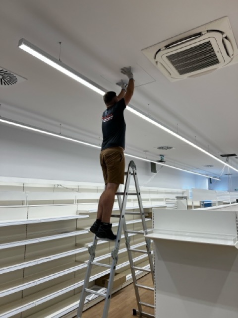 Mężczyzna w rękawicach ochronnych na aluminiowej drabinie, manipulujący przy panelu sufitowym w pomieszczeniu z pustymi regałami sklepowymi, widoczny klimatyzator kasetonowy i oświetlenie liniowe.
