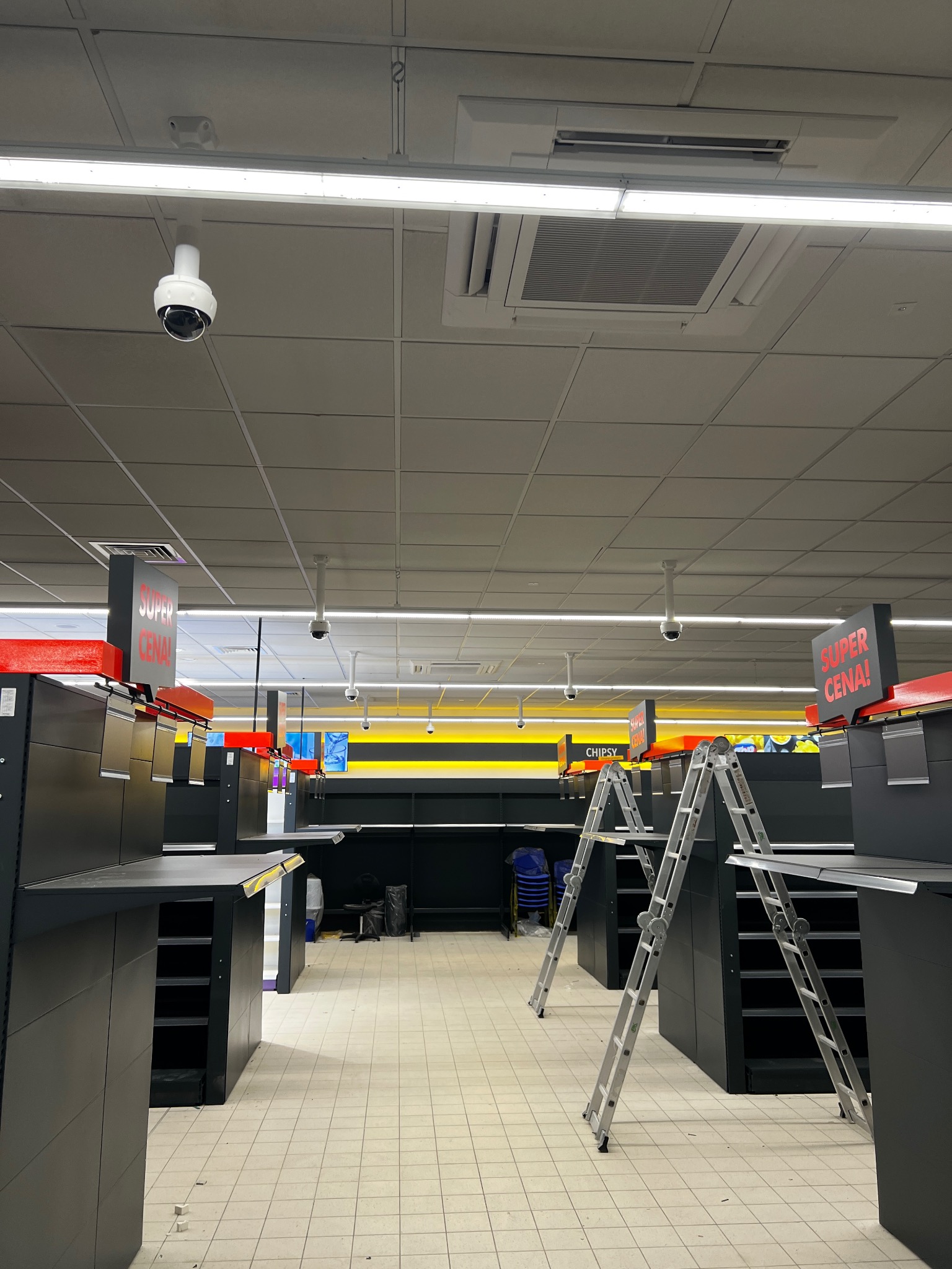 Sklep z pustymi półkami, gdzie na suficie zamontowane są kamery przemysłowe, a na podłodze stoją dwie aluminiowe drabiny.