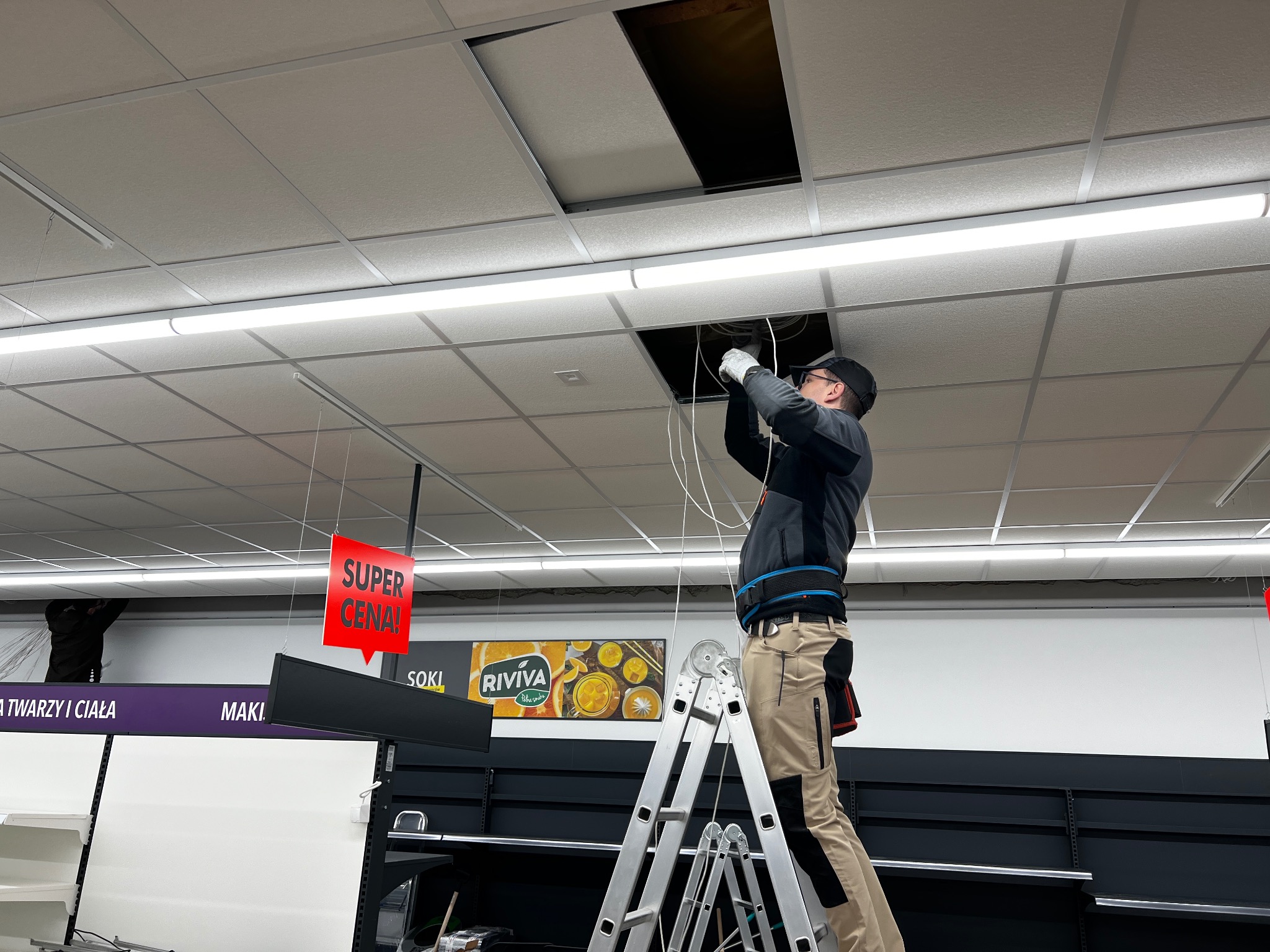 Pracownik na drabinie w sklepie, instalujący lub naprawiający okablowanie w suficie podwieszanym, z widocznym otworem rewizyjnym i elementami oświetlenia.