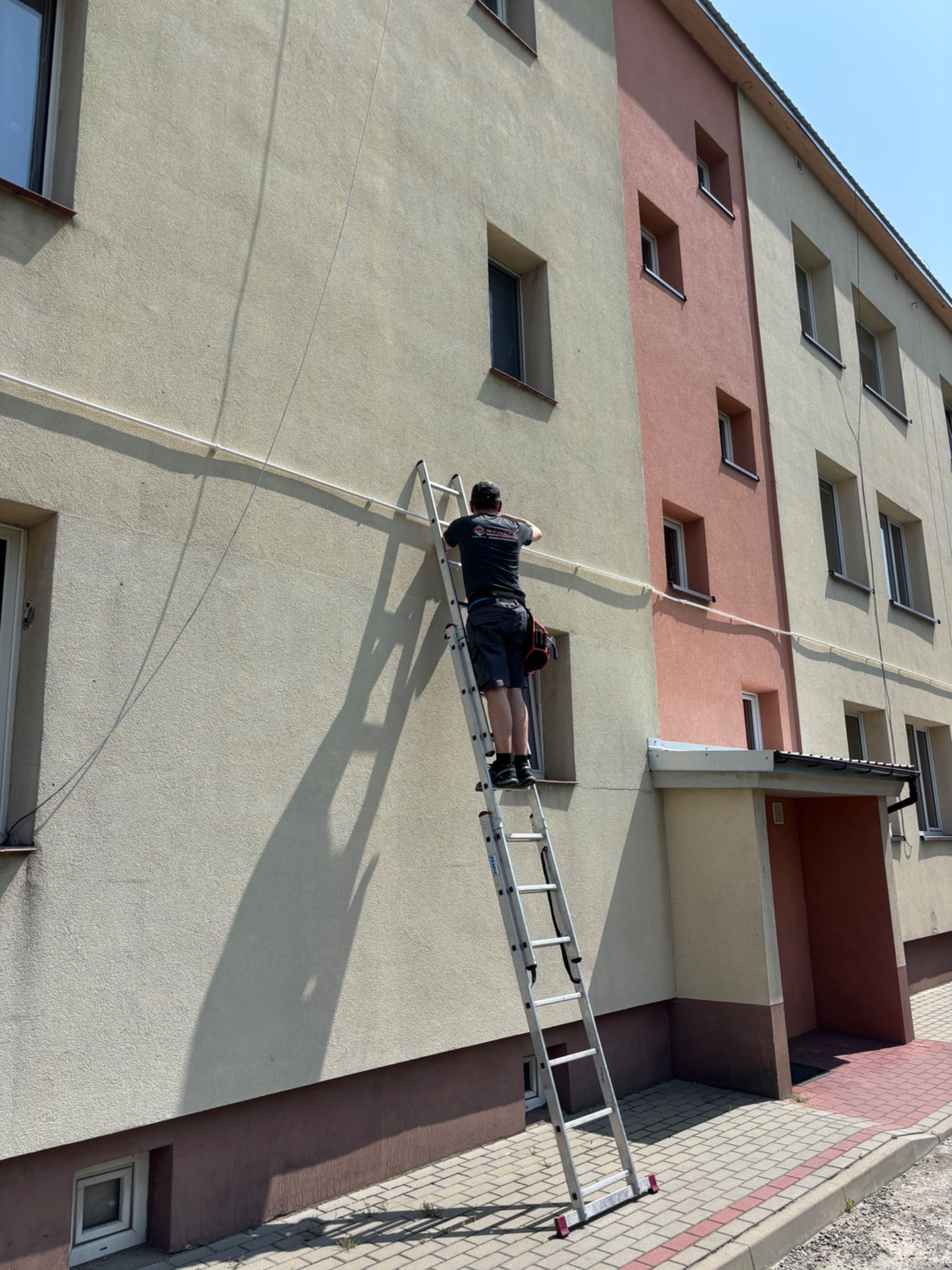 Instalator na drabinie montuje przewód na elewacji budynku. Widoczna torba z narzędziami, budynek wielorodzinny. Słoneczny dzień, cień osoby na ścianie.