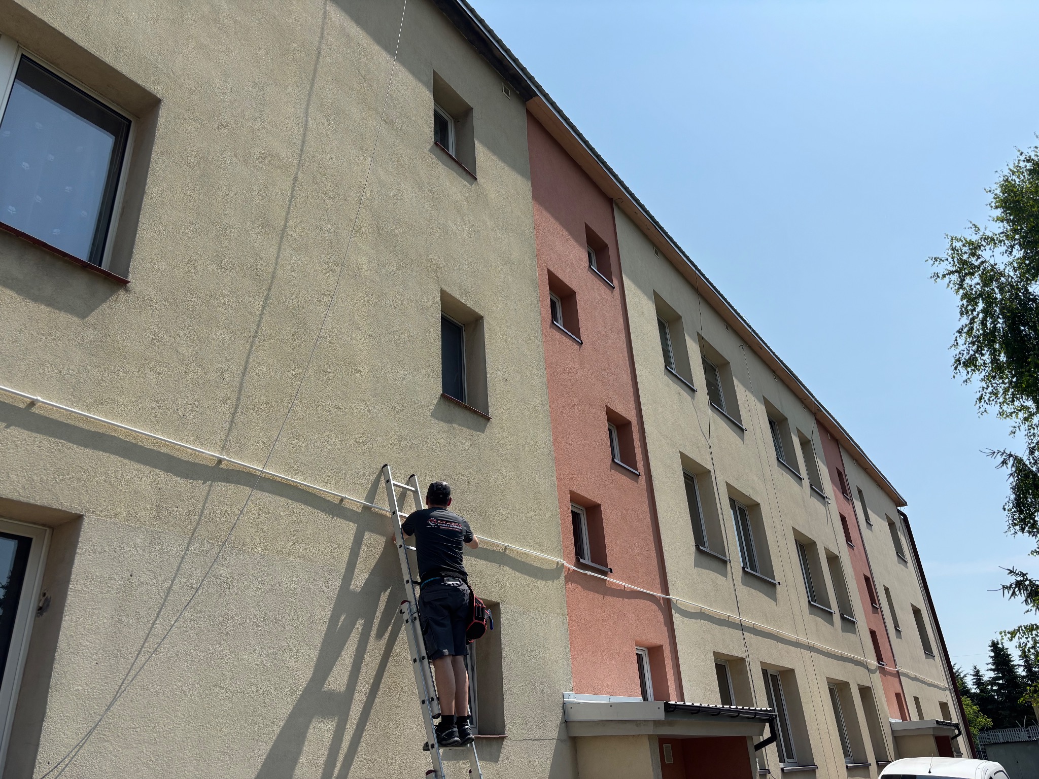 Instalator na drabinie montuje białe przewody na elewacji budynku wielorodzinnego, widoczne okna i fragment drzewa. Praca na wysokości w słoneczny dzień.