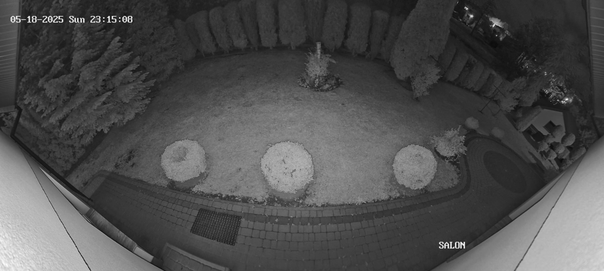 Nocny widok z kamery monitoringu na posesję z trawnikiem, krzewami i domem. Widoczne data i godzina nagrania w lewym górnym rogu.