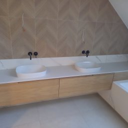 STUDIO RS MEBLE NA WYMIAR - Nowoczesna łazienka z dwoma białymi umywalkami nablatowymi, zamontowanymi na drewnianej szafce z szufladami, jasne płytki na ścianie ułożone w jodełkę, widoczne niedokończone podłączenia hydrauliczne.