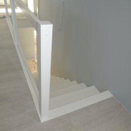 STUDIO RS MEBLE NA WYMIAR - Nowoczesne białe schody z drewnianymi stopniami i białą balustradą z elementami szklanymi, widok z góry, w tle szara ściana i wystające przewody.