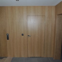 STUDIO RS MEBLE NA WYMIAR - Ściana z ukrytymi drzwiami z drewna, wkomponowana w drewnianą boazerię, z widocznymi włącznikami światła i fragmentem podłogi.