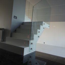 STUDIO RS MEBLE NA WYMIAR - Nowoczesne, białe schody wspornikowe z szklaną balustradą, mocowaną za pomocą srebrnych uchwytów, na tle białej ściany i ciemnej, marmurowej podłogi.