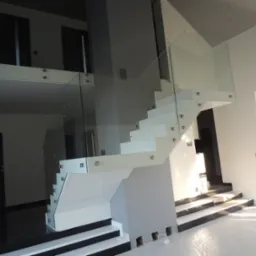 Nowoczesne, białe schody z przezroczystą szklaną balustradą, mocowaną za pomocą srebrnych uchwyt&oacute;w, w jasnym wnętrzu z czarnymi akcentami na podłodze, widoczne otwory w ścianie na dole schod&oacute;w.