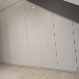 STUDIO RS MEBLE NA WYMIAR - Biała szafa wnękowa z połyskiem, dopasowana do skośnego sufitu, z widoczną podłogą z paneli w jasnym odcieniu drewna i fragmentem ciemnej ściany po lewej stronie.