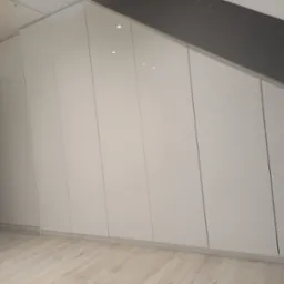 Biała szafa wnękowa z połyskiem, dopasowana do skośnego sufitu, z widoczną podłogą z paneli w jasnym odcieniu drewna i fragmentem ciemnej ściany po lewej stronie.