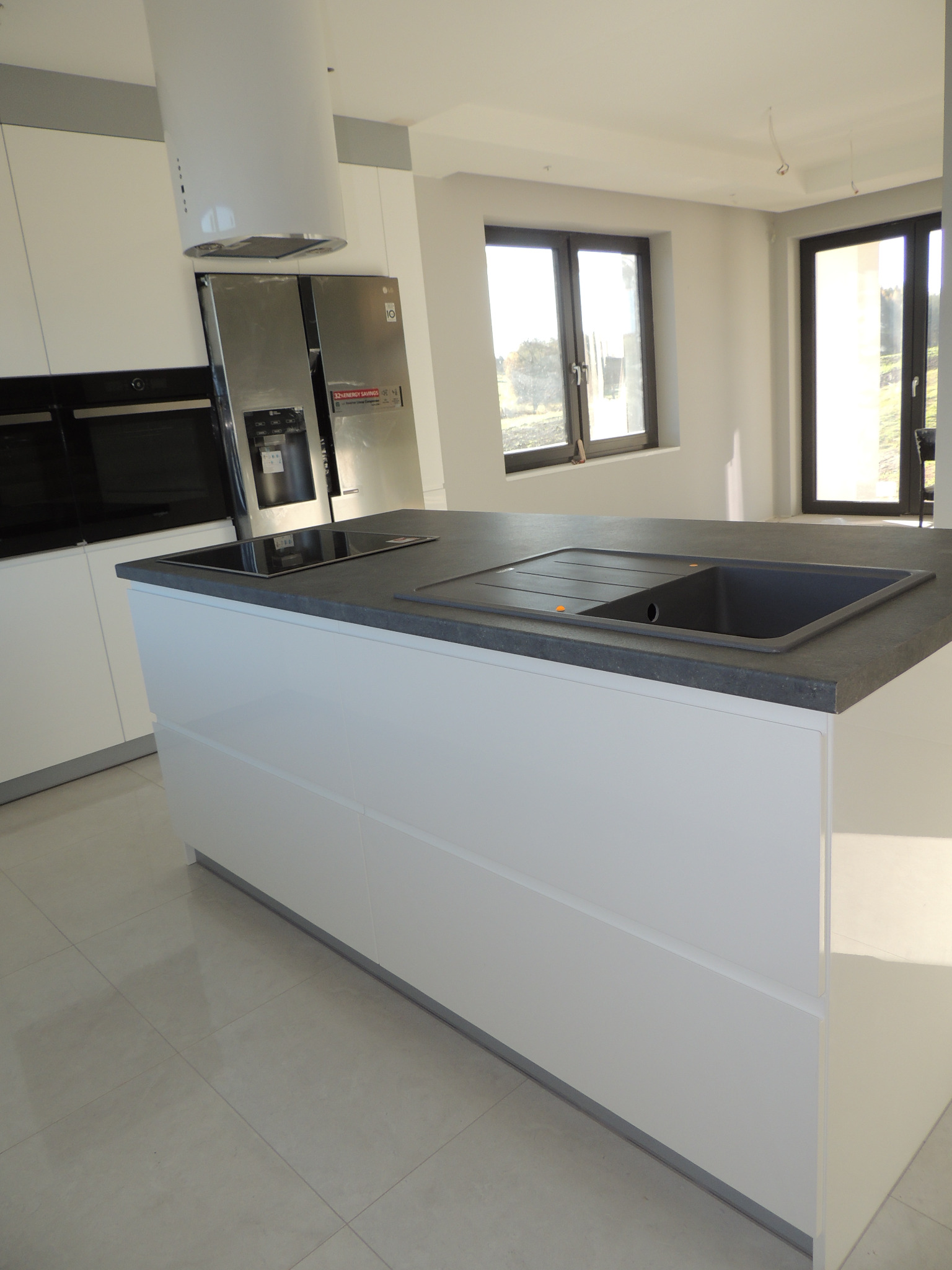 Nowoczesna, biała wyspa kuchenna z szarym blatem, wbudowanym zlewem i płytą indukcyjną. W tle widoczna zabudowa kuchenna z piekarnikiem i lodówką oraz okna w jasnym wnętrzu.