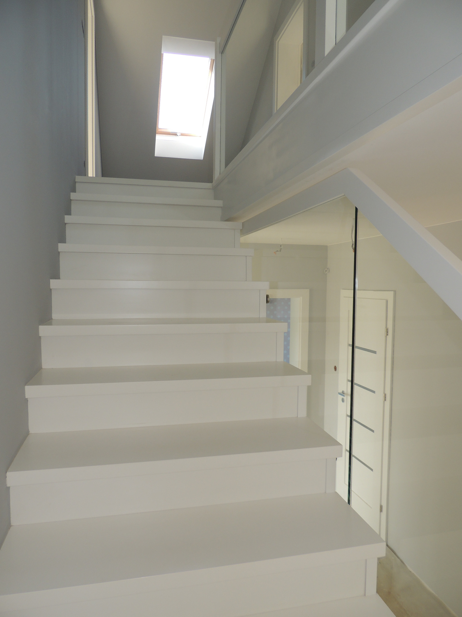 Białe schody prowadzące na poddasze z oknem dachowym, minimalistyczny design, szklana balustrada, białe drzwi w tle.
