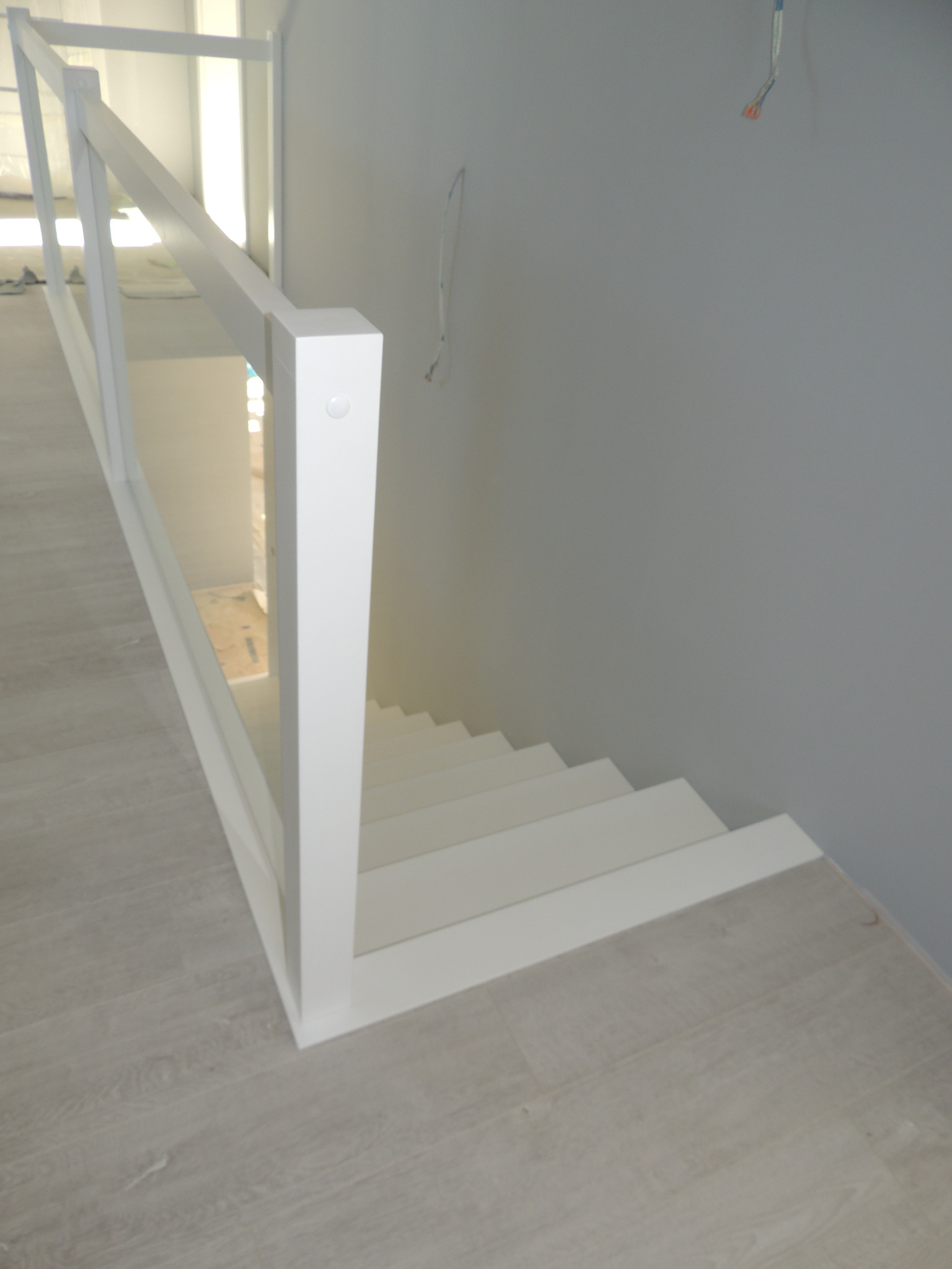 Nowoczesne białe schody z drewnianymi stopniami i białą balustradą z elementami szklanymi, widok z góry, w tle szara ściana i wystające przewody.