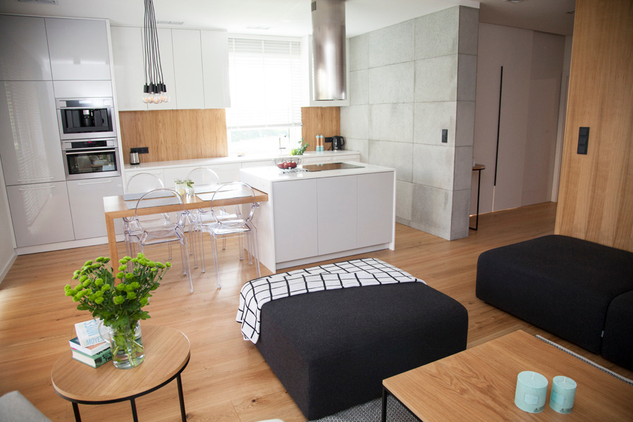 Nowoczesna kuchnia z białymi szafkami na wysoki połysk, drewnianym panelem nad blatem, wyspą kuchenną i przezroczystymi krzesłami, uzupełniona elementami dekoracyjnymi w salonie.