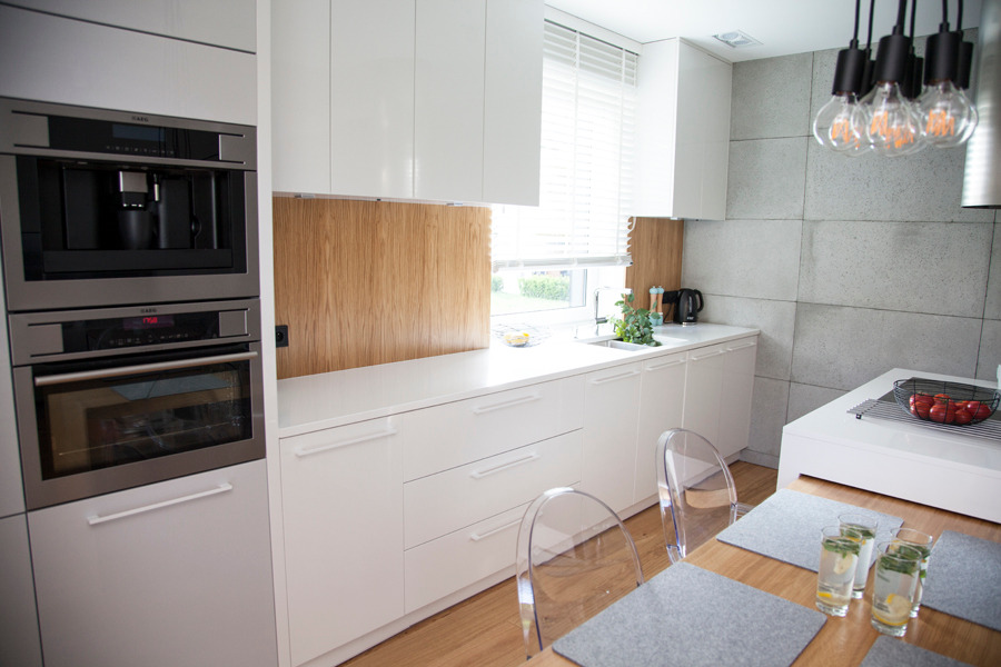 Nowoczesna, jasna kuchnia z białymi frontami, drewnianym panelem ściennym i wbudowanym sprzętem AGD, widok na stół z przezroczystymi krzesłami.