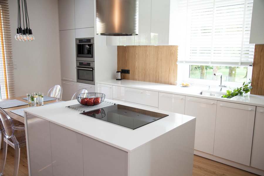 Nowoczesna kuchnia z wyspą, białymi lakierowanymi frontami, drewnianym panelem na ścianie i wiszącymi lampami z żarówkami Edisona.