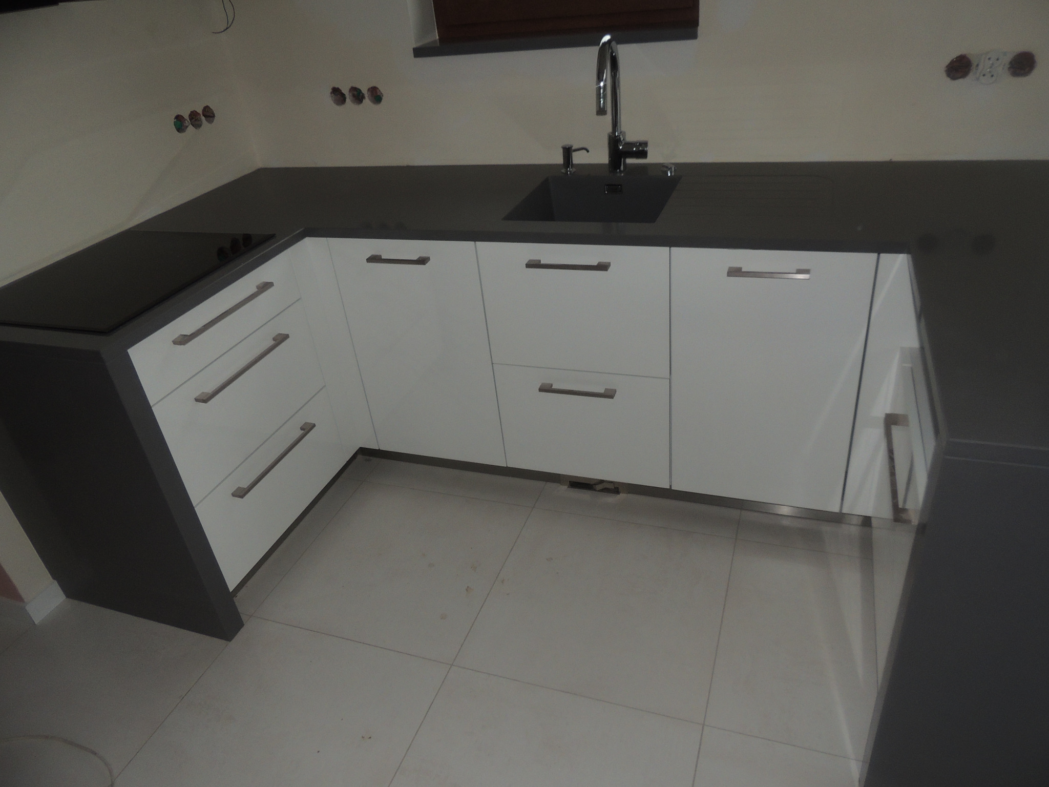 Nowoczesna kuchnia z białymi frontami szafek i ciemnym blatem, widoczny zlew z baterią i płyta indukcyjna, brakujące wykończenie ściany w tle.