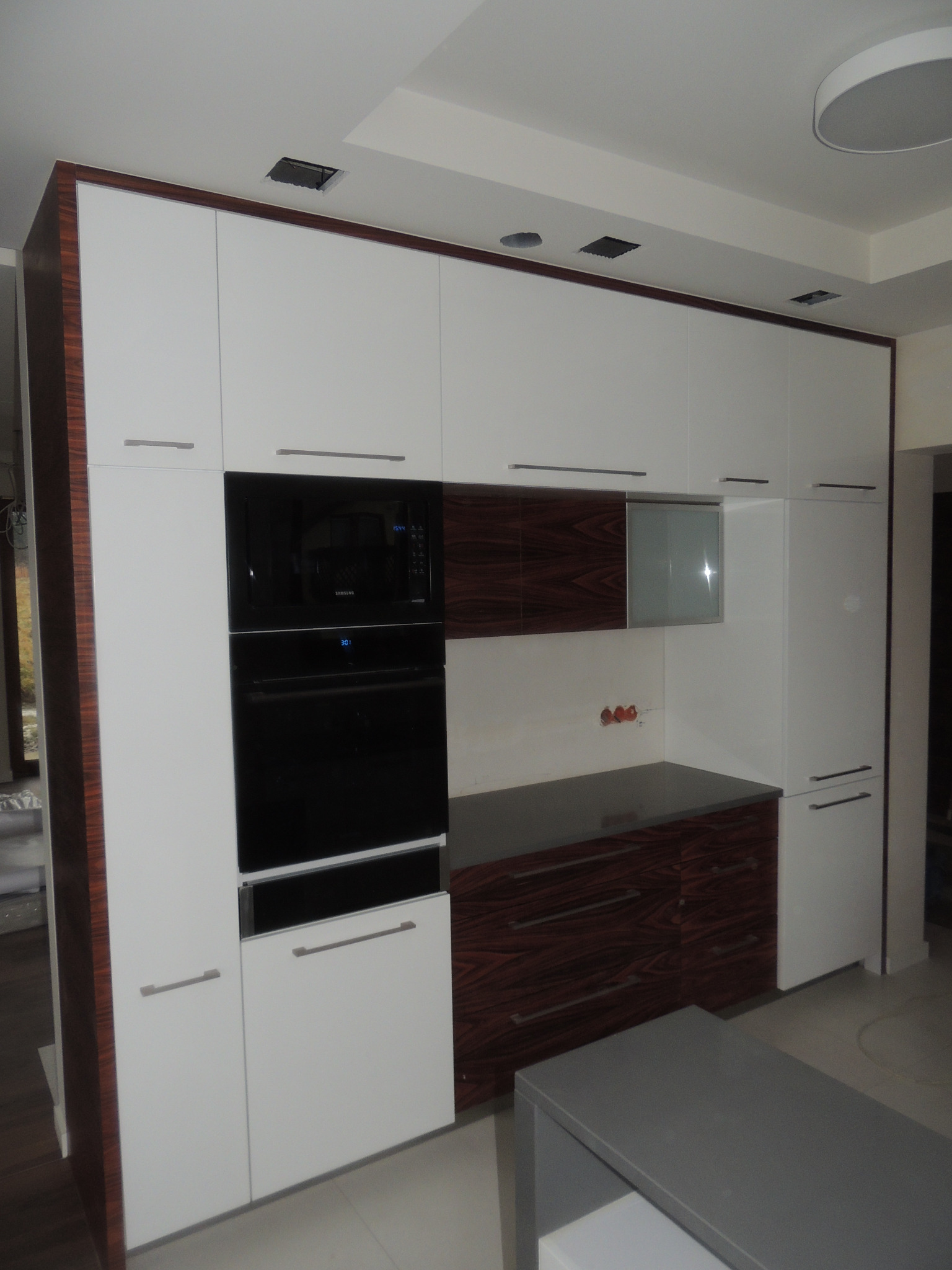 Nowoczesna zabudowa kuchenna z białymi frontami i elementami w odcieniu ciemnego drewna, z wbudowanym piekarnikiem i mikrofalówką Samsung, oświetleniem halogenowym w suficie.