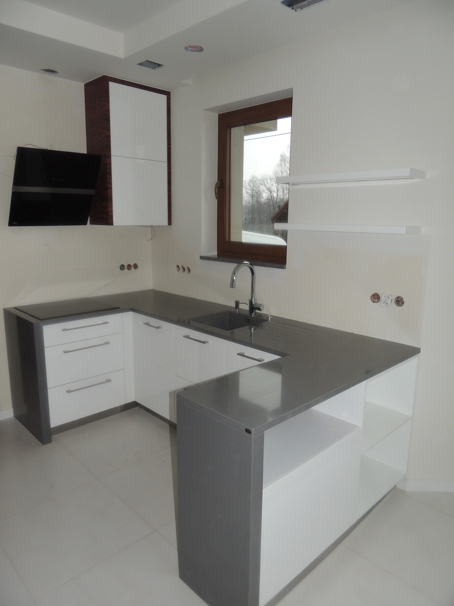 Nowoczesna kuchnia z białymi frontami i szarym blatem, widok na zlew i okno, minimalistyczny design, gładkie powierzchnie i brak uchwytów, czarny okap.