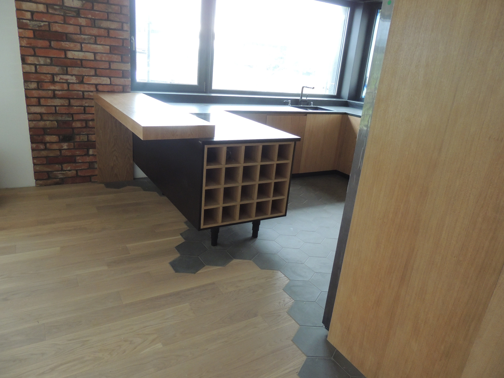 Kuchnia z wyspą z wbudowanym regałem na wino, ceglana ściana w tle, podłoga z drewnianych paneli przechodząca w heksagonalne płytki.