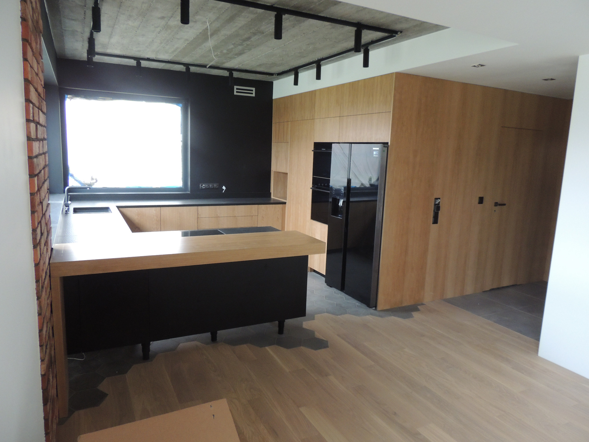 Nowoczesna kuchnia z drewnianymi frontami, czarnym blatem i wyspą z czarnymi szafkami, oświetleniem szynowym i ceglanym wykończeniem ściany.