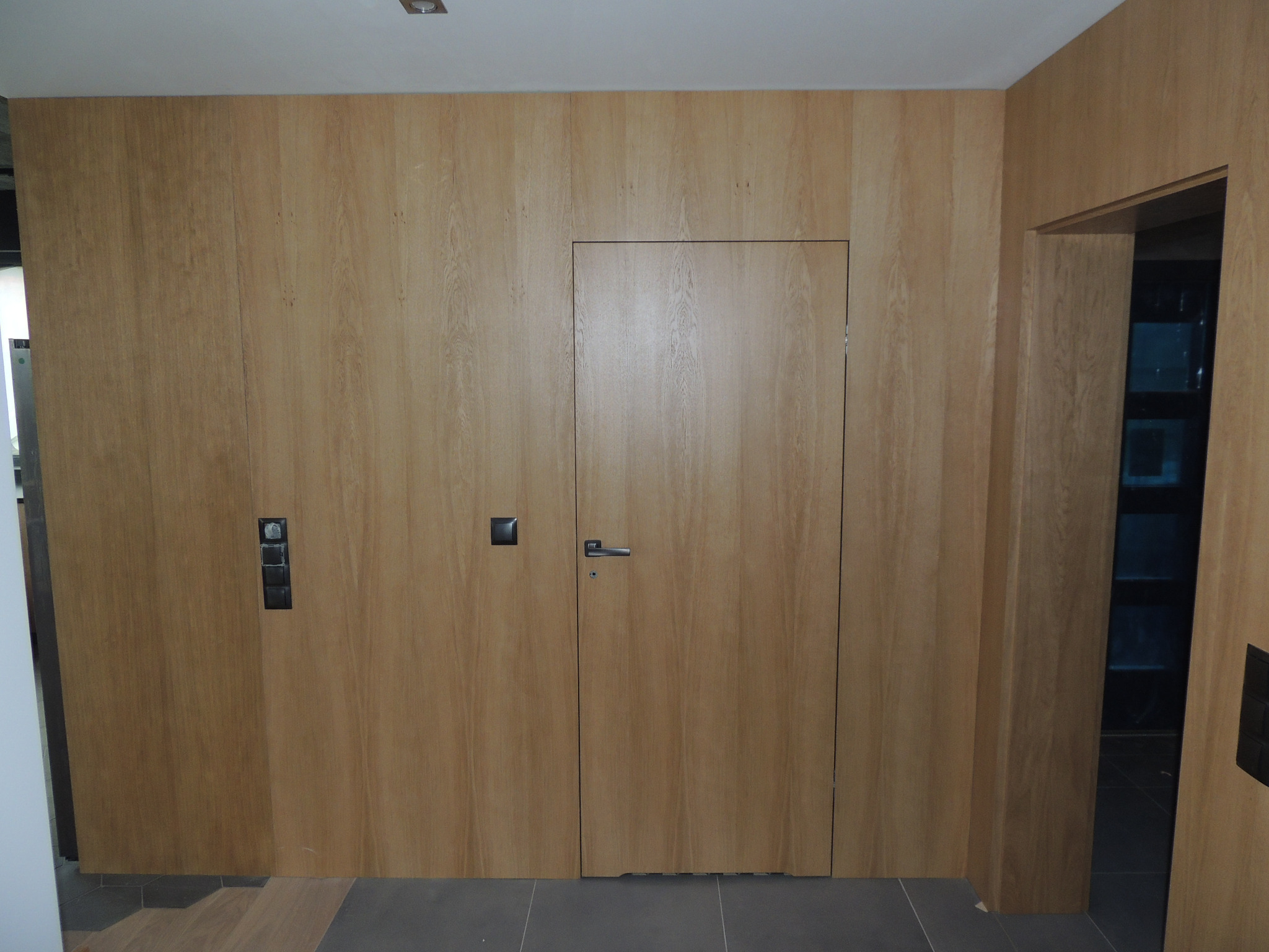 Ściana z ukrytymi drzwiami z drewna, wkomponowana w drewnianą boazerię, z widocznymi włącznikami światła i fragmentem podłogi.