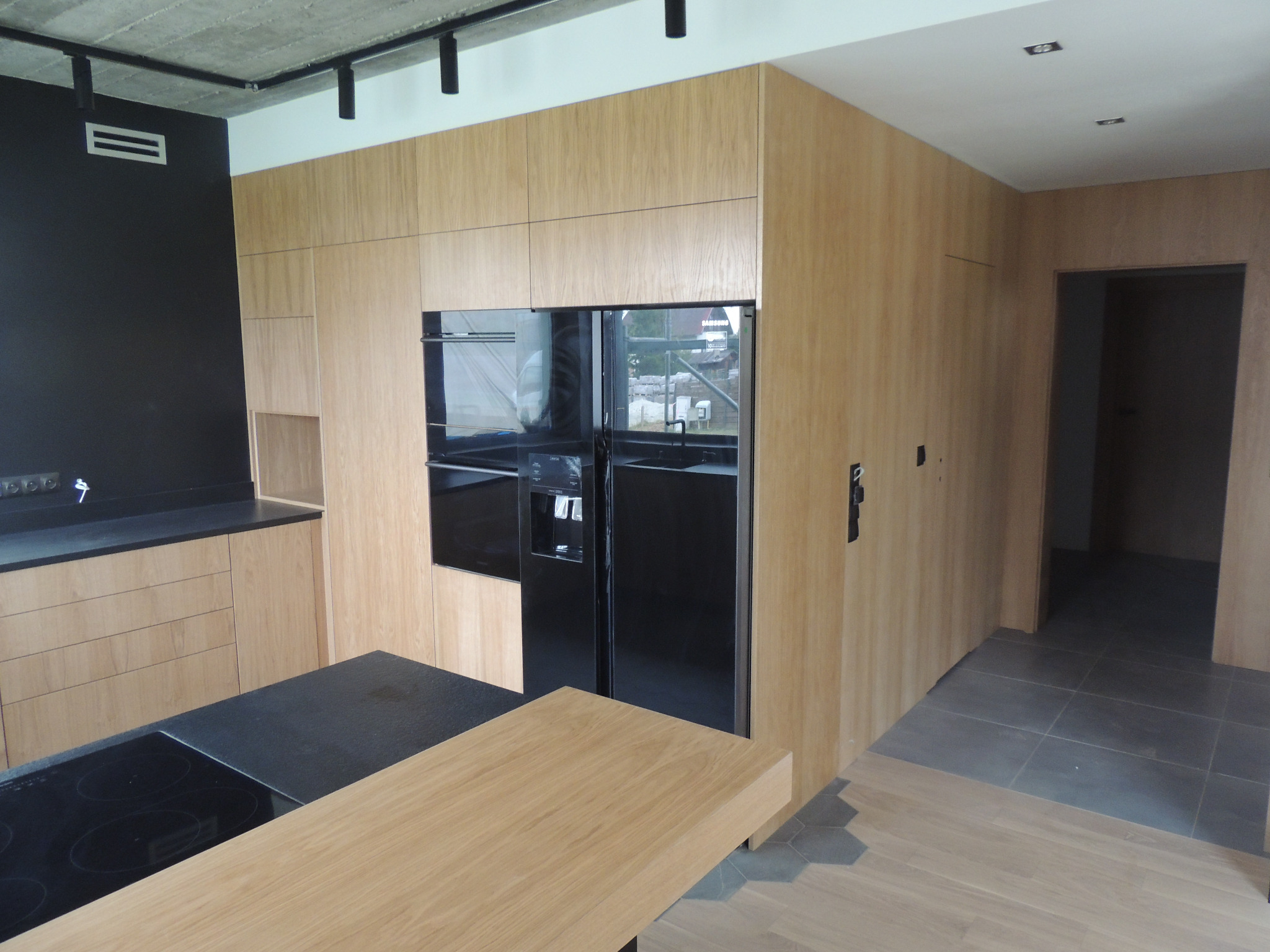 Nowoczesna kuchnia z drewnianymi frontami, czarnym blatem i zabudowanym sprzętem AGD, widok na wyspę kuchenną z płytą indukcyjną i drewnianym blatem, surowy betonowy sufit z oświetleniem szynowym.