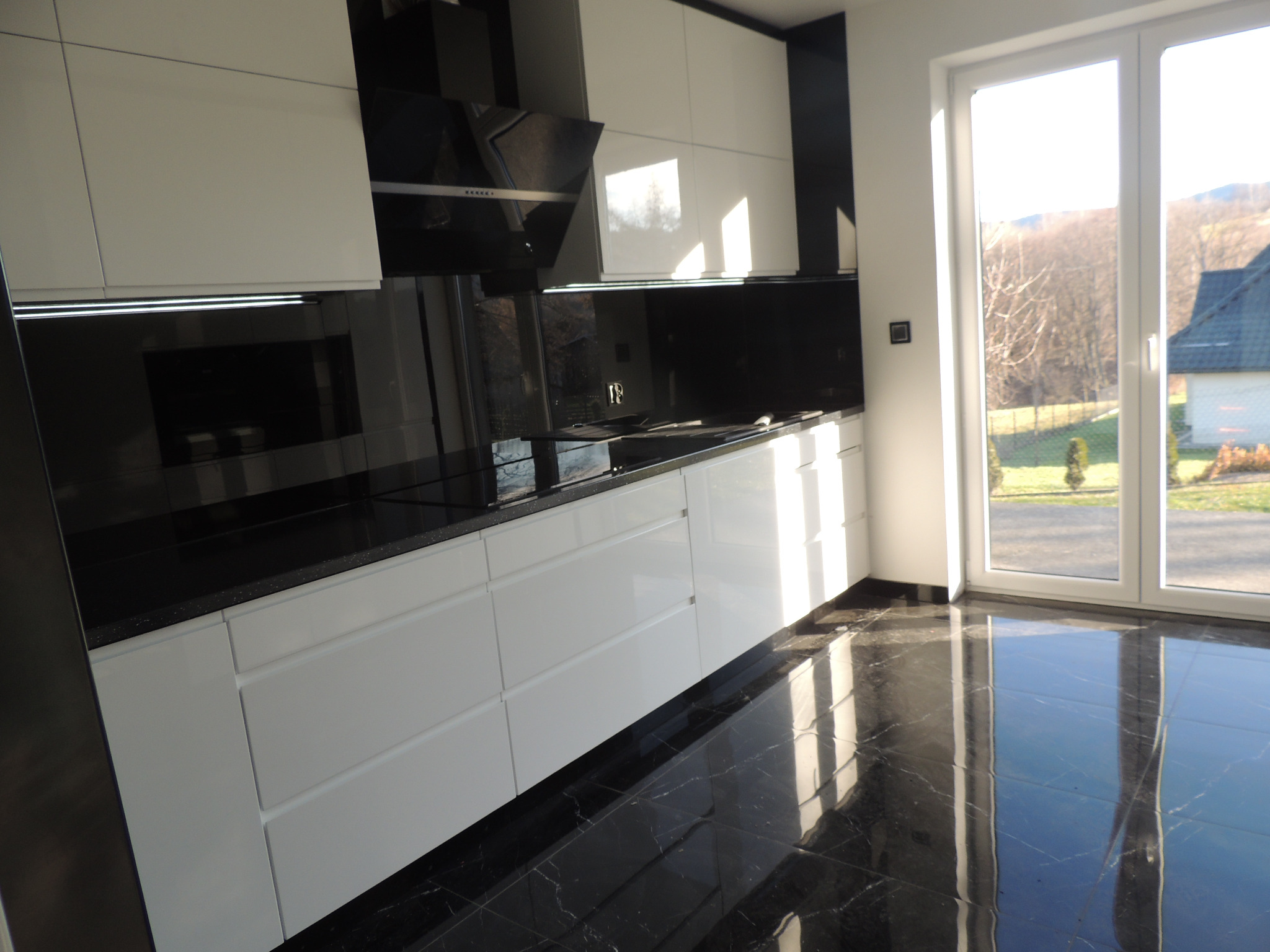 Nowoczesna kuchnia z białymi szafkami i czarnym blatem, odbijająca światło w polerowanej, marmurkowej podłodze, z widokiem na zewnątrz przez okno.