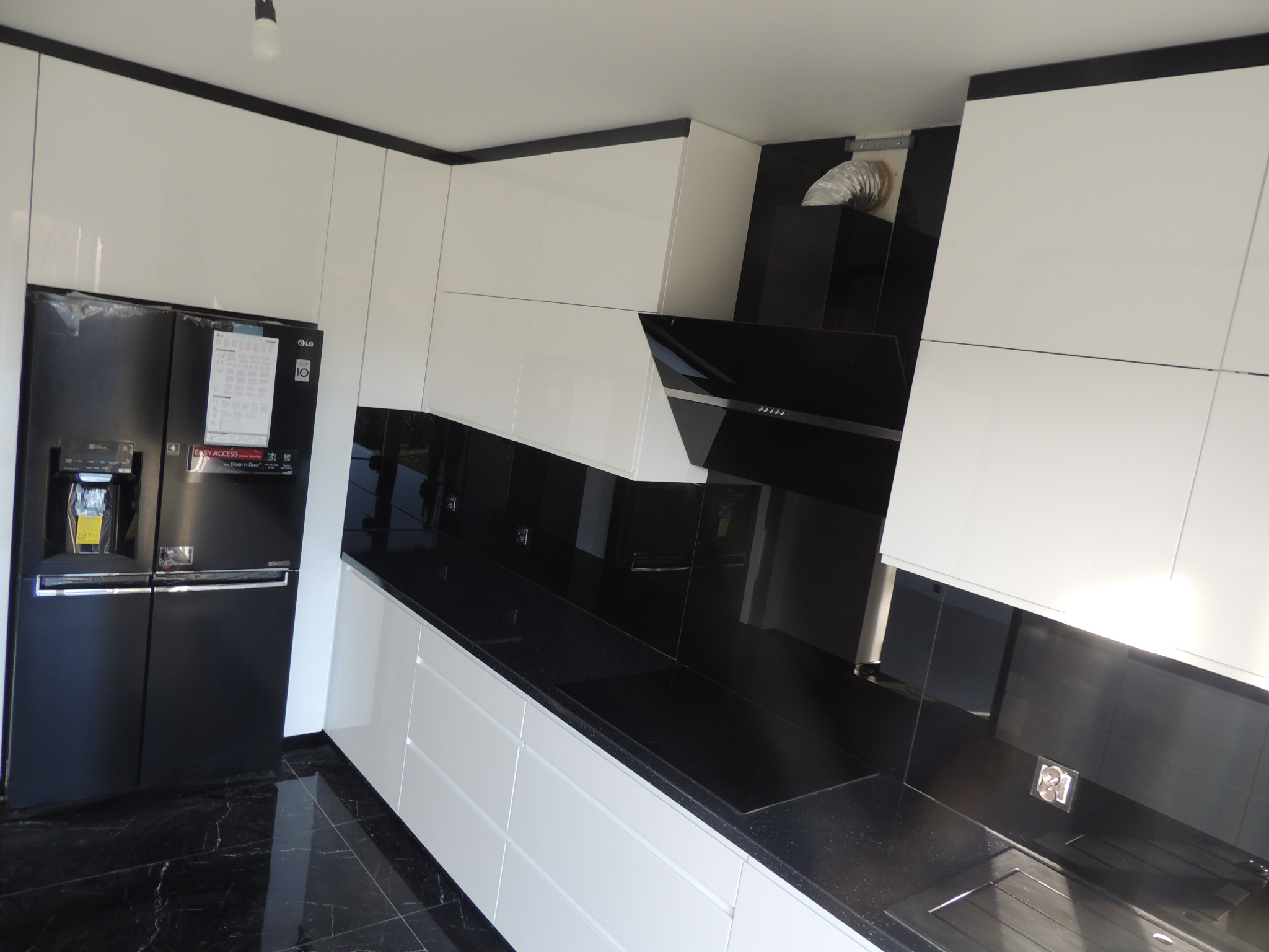 Nowoczesna kuchnia z białymi szafkami i czarnym blatem, widoczny czarny dwudrzwiowy lodówka LG i marmurowa podłoga.