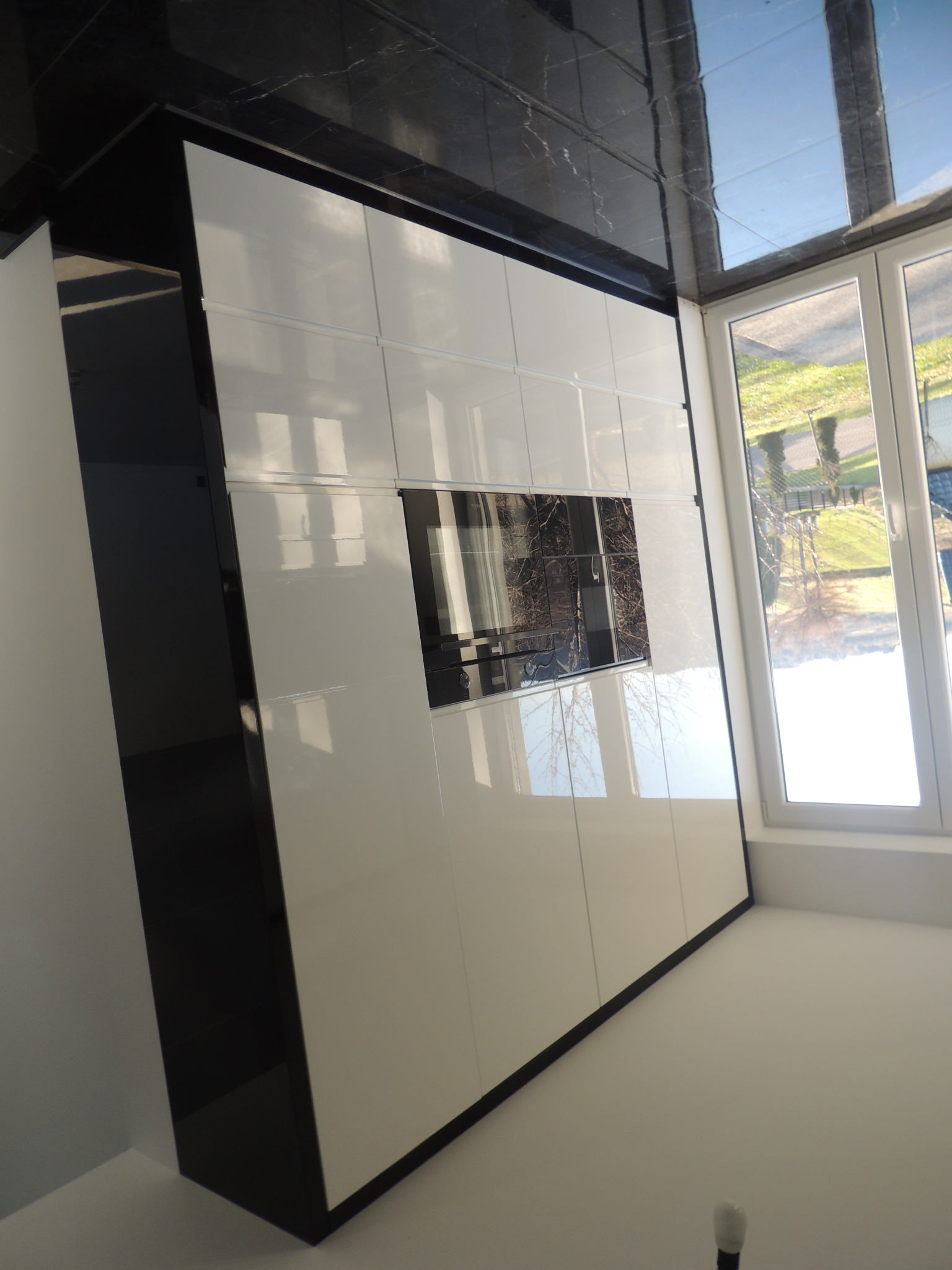 Biała szafa wnękowa z połyskiem, czarne obramowanie, zabudowa pod sufit, widoczne okno z odbiciem drzew