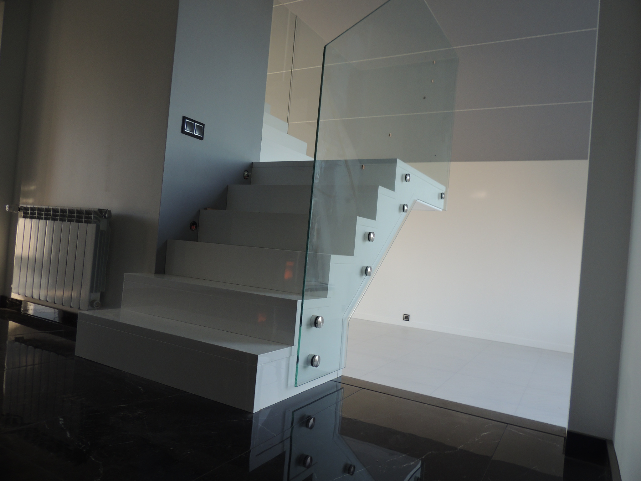 Nowoczesne, białe schody wspornikowe z szklaną balustradą, mocowaną za pomocą srebrnych uchwytów, na tle białej ściany i ciemnej, marmurowej podłogi.