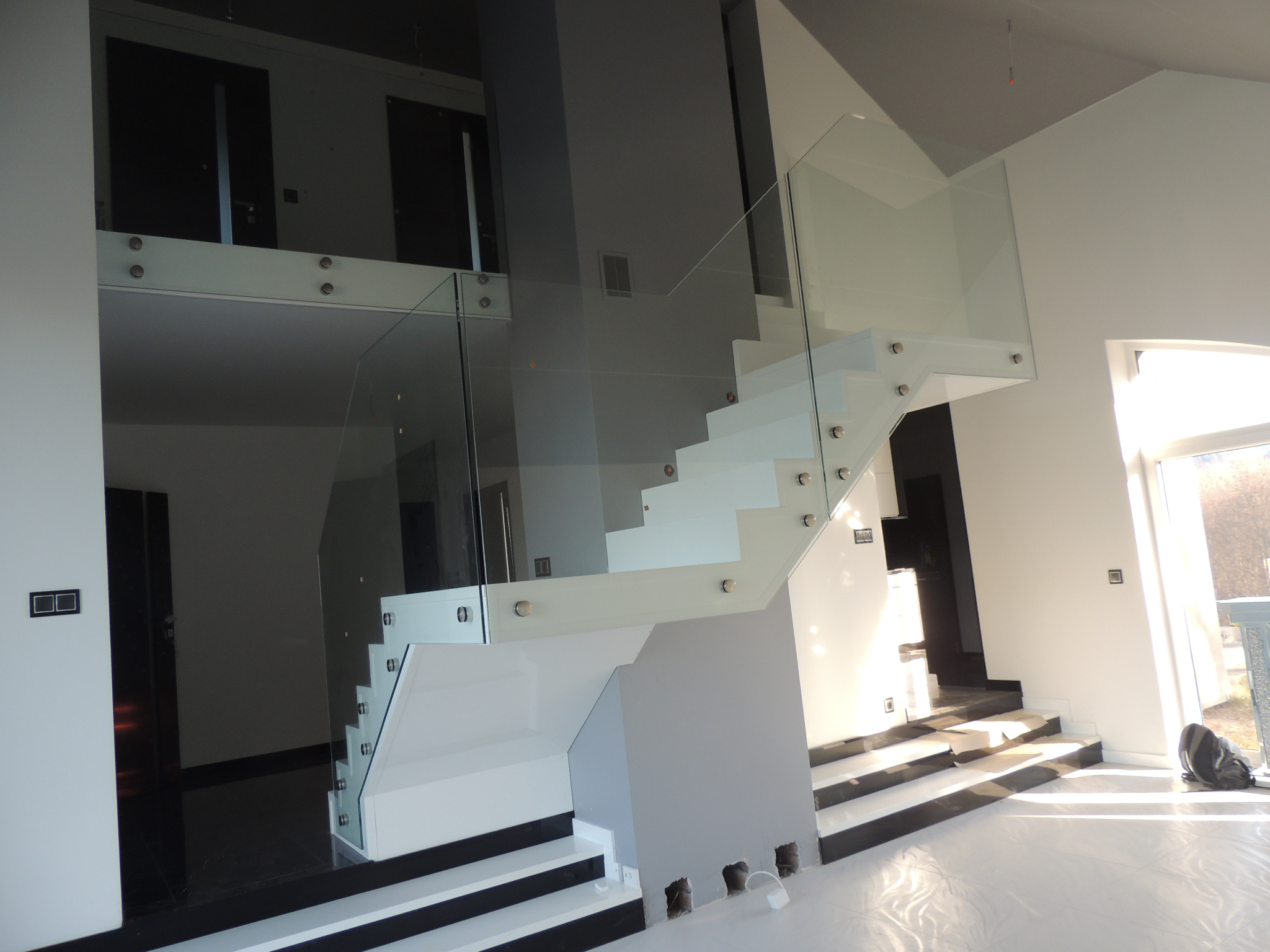 Nowoczesne, białe schody z przezroczystą szklaną balustradą, mocowaną za pomocą srebrnych uchwytów, w jasnym wnętrzu z czarnymi akcentami na podłodze, widoczne otwory w ścianie na dole schodów.