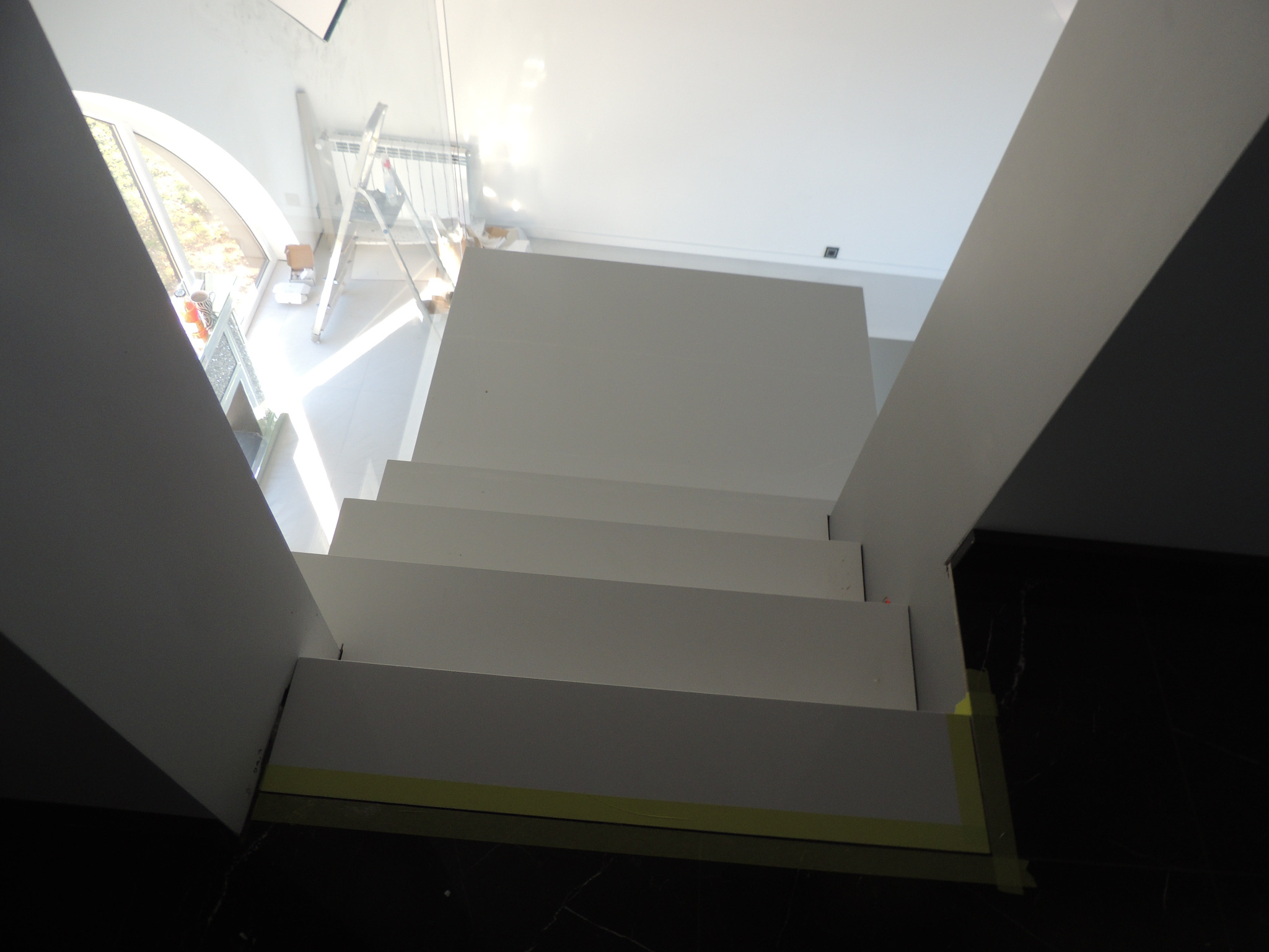 Nowoczesne, białe schody z widocznymi taśmami malarskimi zabezpieczającymi podłogę, w tle okno łukowe i drabina malarska.