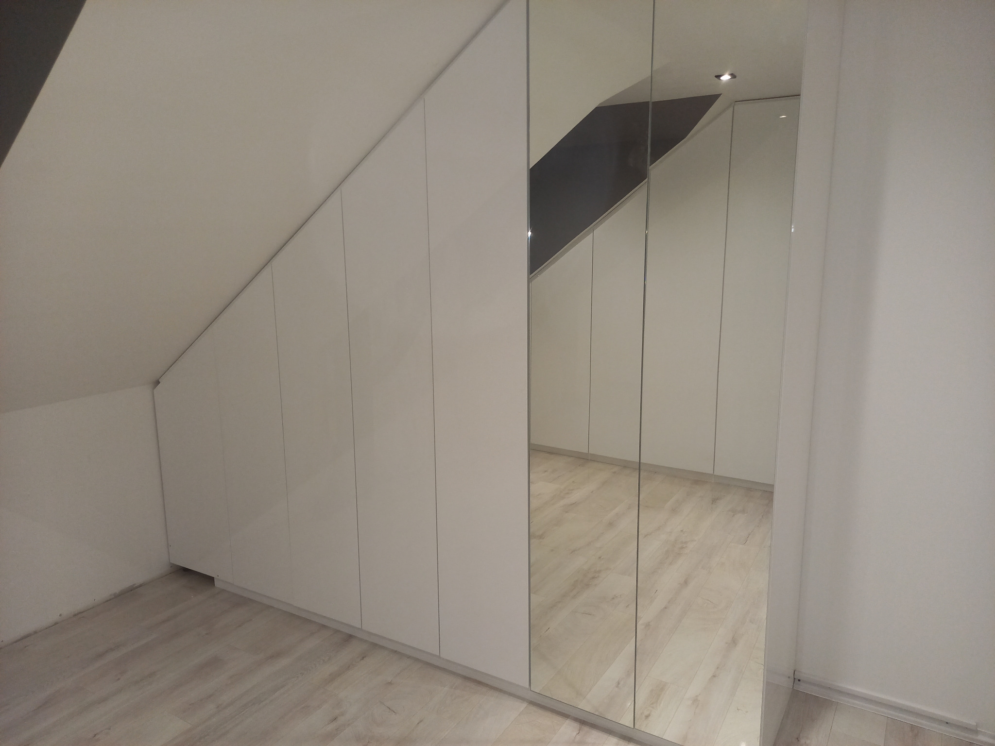 Biała szafa wnękowa z lustrzanymi drzwiami, dopasowana do skosu poddasza, widoczne odbicie podłogi w lustrze.
