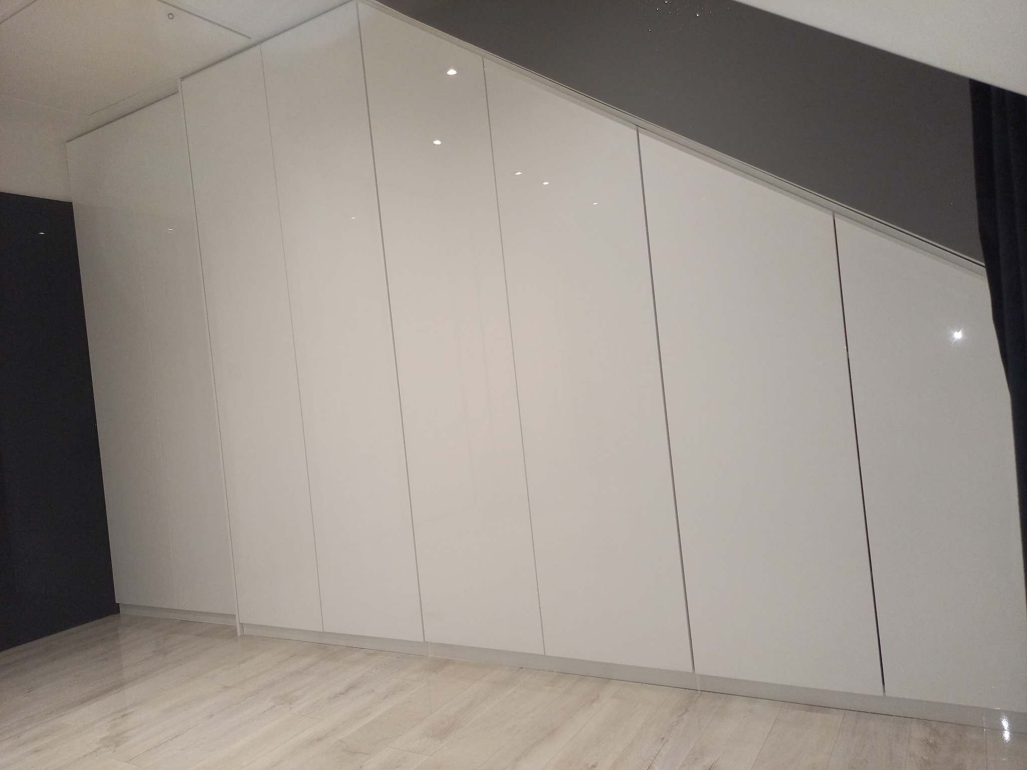 Biała szafa wnękowa z połyskiem, dopasowana do skośnego sufitu, z widoczną podłogą z paneli w jasnym odcieniu drewna i fragmentem ciemnej ściany po lewej stronie.