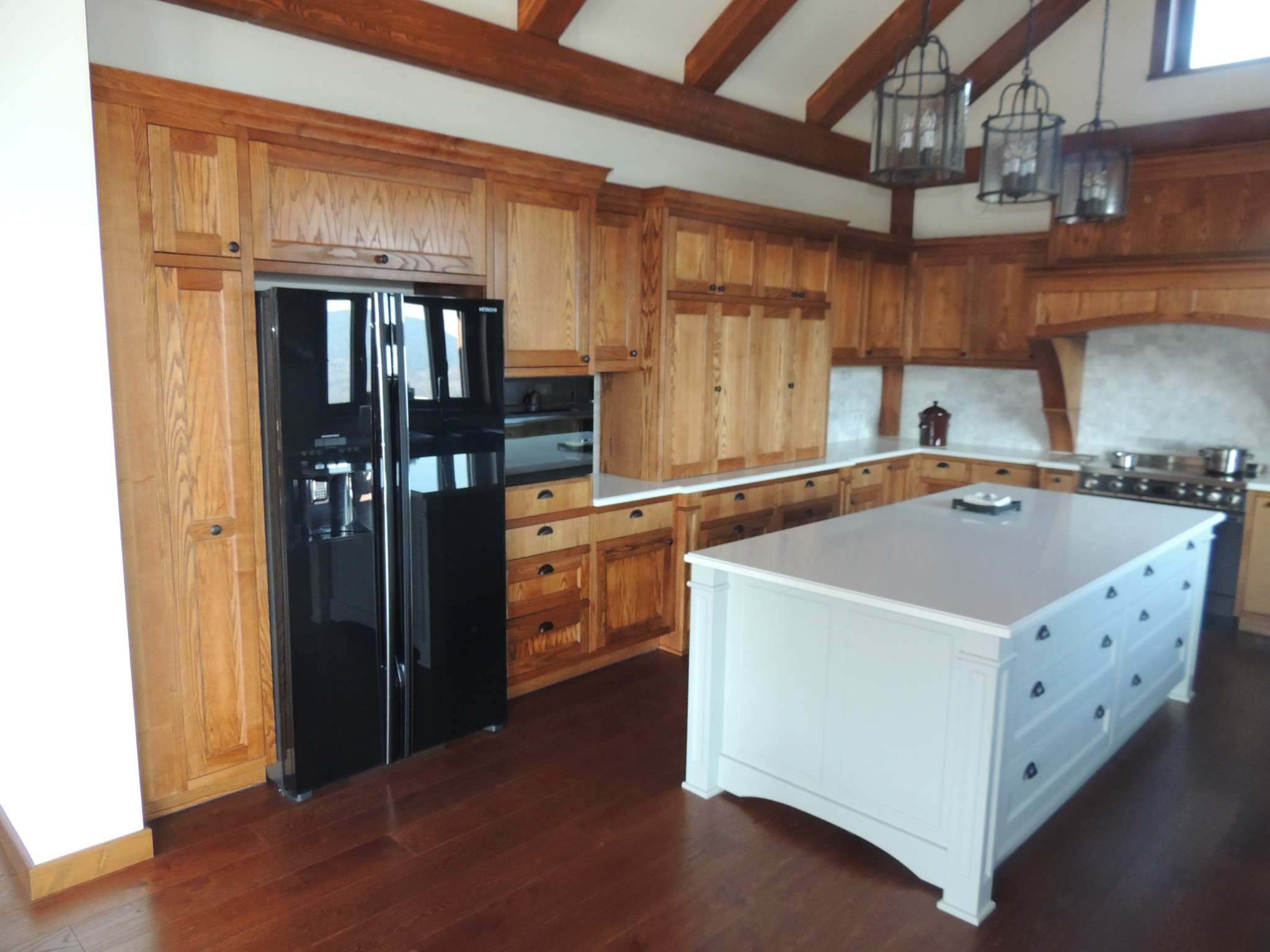 Jasna, przestronna kuchnia z drewnianymi szafkami, czarną lodówką i wyspą z szufladami, oświetlona wiszącymi lampami w stylu industrialnym.